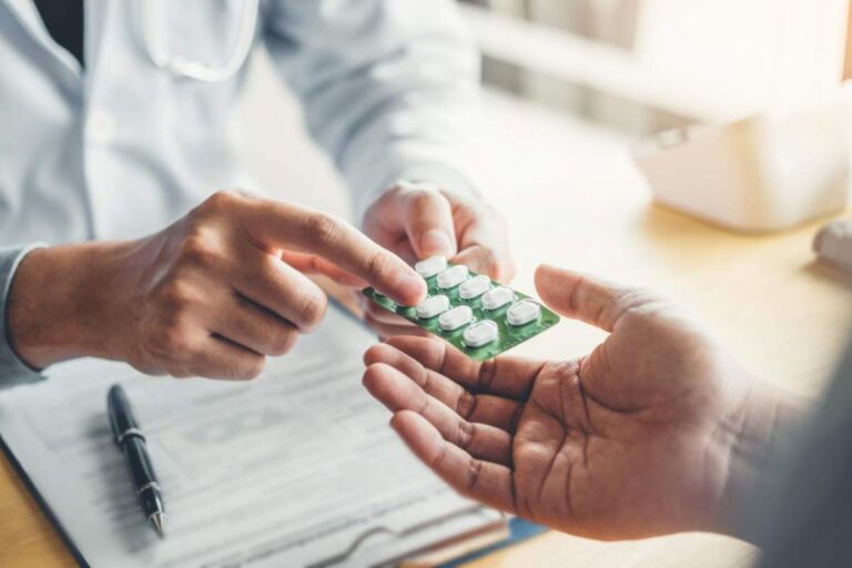 A hand holds several colorful pills above a wooden kitchen table, showing their variety and differences.