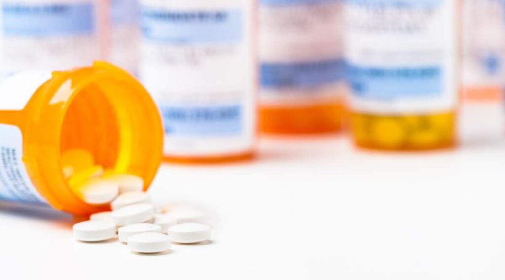 A hand holding two prescription pill bottles with capsules and tablets of different shapes and colors spread on a table.