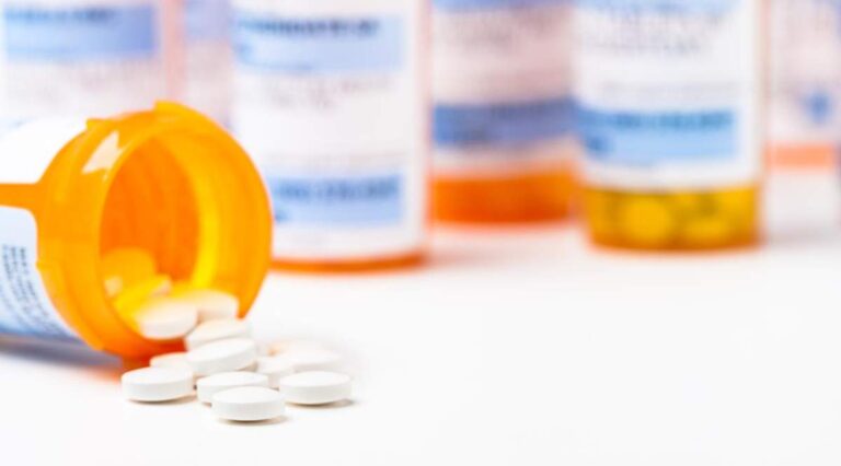 A hand holding two prescription pill bottles with capsules and tablets of different shapes and colors spread on a table.